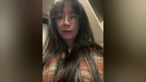 Video of a woman with long black hair, wearing glasses and a patterned orange shirt, taken indoors, blurred background.