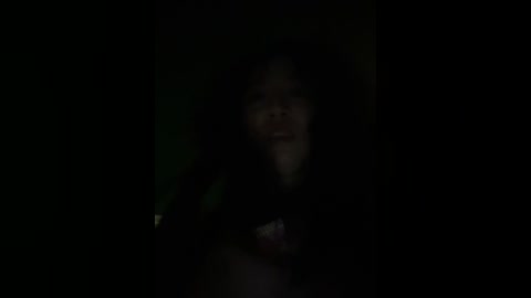 A dimly lit video of a young girl with long, dark hair, partially obscured by shadow, smiling warmly. The background is dark and indistinct, emphasizing the girl's face.
