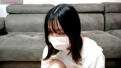 Video of an East Asian woman with long, straight black hair, wearing a white mask, kneeling on a beige couch in a minimalist living room.