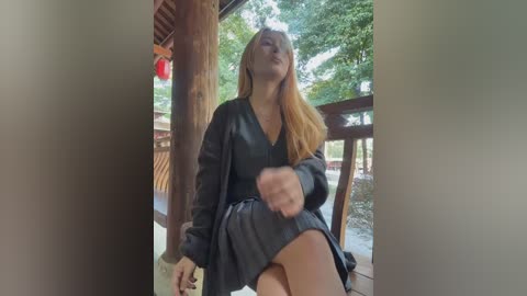 A video of a young woman with long blonde hair in a black dress, holding a milk bottle, sitting on a wooden porch with trees and a red lantern in the background.