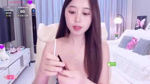 A video of a young Asian woman with long brown hair, wearing a white top, applying makeup in a modern bedroom with a bed, nightstand, and lamp.