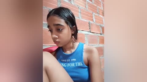 Video of a young Southeast Asian girl with light brown skin, long black hair, wearing a blue ribbed tank top with white text, sitting against a red brick wall.