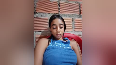 Video of a young woman with medium skin tone and long black hair, wearing a blue ribbed tank top with white text, sitting against a red-brick wall, looking thoughtful.