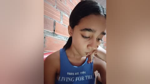 Video of a young girl with medium brown skin, straight dark hair, and light blue sleeveless top, sitting on a red chair, gazing thoughtfully at her hand. Brick wall background.