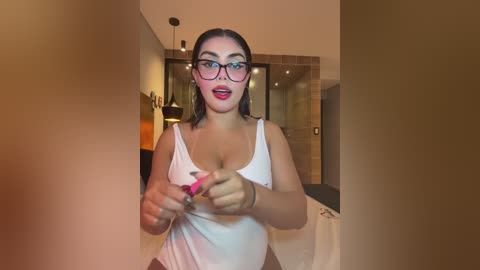 Video of a young woman with medium skin tone and shoulder-length brown hair, wearing glasses, a white tank top, and pink lipstick. She's pulling down her top in a bathroom with beige tiles, a shower, and a sink.
