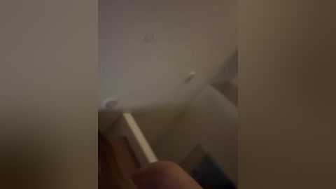 A dimly lit video of a hand holding a white candlestick, partially obscured by shadows, in a dimly lit room with a beige wall and indistinct furniture.