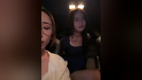 Video of two women in a dimly-lit car, one with glasses, the other with long hair and a dark dress, looking forward, blurred background.