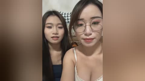 Video of two young Asian women with long black hair, wearing glasses and light-colored tops, sitting closely together, indoors with a checkered patterned wall behind them.