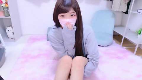 A video of an Asian woman in a light blue jacket, wearing a pink face mask, sitting on a pink, fluffy bed in a minimalist room with white walls and light blue furniture.