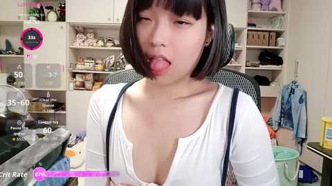 A video of an East Asian woman with a bob haircut, sticking out her tongue, wearing a low-cut white top in a cluttered, brightly-lit room.