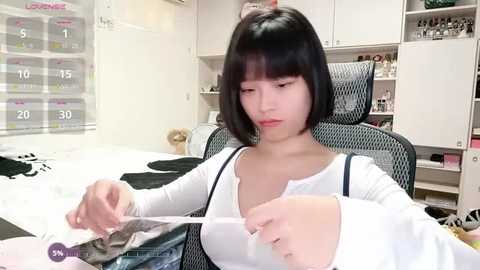 Video of an East Asian woman with short black hair in a white top, seated at a desk with a calendar and various personal items, including makeup and a pair of shoes, in a tidy room.