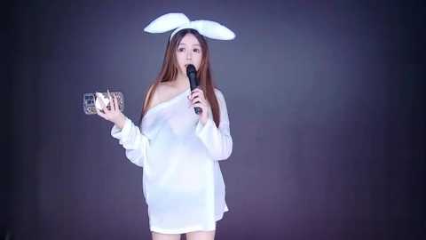Video of an Asian woman with long brown hair, wearing a white off-shoulder dress, rabbit ears headband, holding a microphone and a glittery fan, against a dark background.