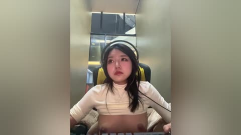 A video of an Asian woman with medium skin tone and long black hair, wearing headphones, sitting in a gaming chair, partially revealing her breasts. The room is dimly lit, with a yellow and black gaming setup in the background.