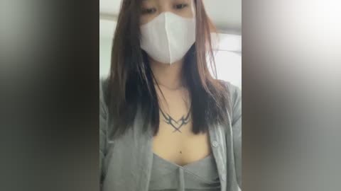 A video of an East Asian woman with long, straight, brown hair, wearing a white surgical mask, a gray shirt with a low neckline revealing cleavage, and a necklace. Background is blurred.