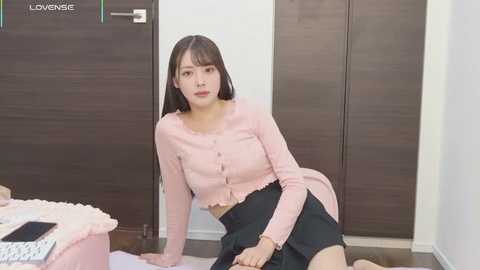 Video of an East Asian woman with straight, dark hair, wearing a pink cardigan and black skirt, kneeling on a floor, in a modern, minimalist bedroom with wooden doors.