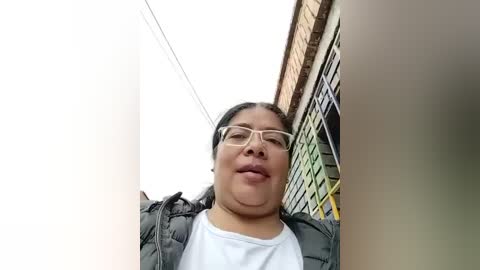 Video of a middle-aged Asian woman with glasses, fair skin, and a puffy face, wearing a black puffy jacket over a white shirt, standing in a narrow alleyway with brick buildings and a white wall on either side.