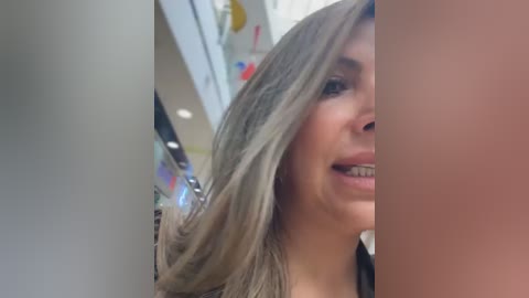 Video of a smiling woman with long, wavy, platinum blonde hair, partially cropped. She has fair skin, and is standing indoors, with a blurred background of a modern, brightly lit building.