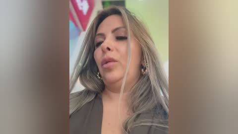 Video of a woman with long, straight, light brown hair, wearing a grey top, looking down, with blurred background showing a red and white sign.