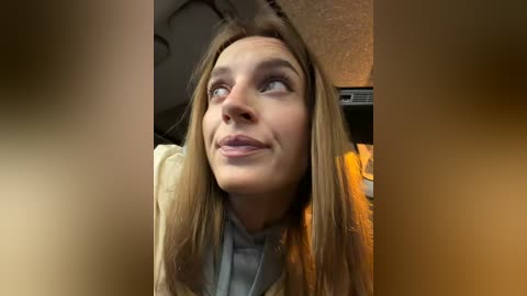 Video of a smiling young woman with long, straight, light brown hair, wearing a beige sweater, in a dimly lit room.