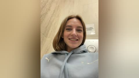 Video of a smiling, light-skinned woman with shoulder-length brown hair, wearing a light blue hoodie with white text, standing in a tiled bathroom with a white wall-mounted light switch.