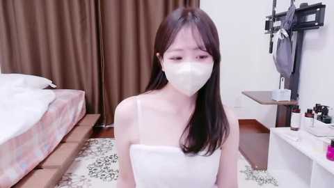 A video of a young Asian woman with long, dark hair, wearing a white face mask and a white towel, sitting on a patterned rug in a sparsely furnished bedroom with a bed, wooden floor, and brown curtains.