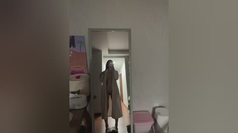 Video of a woman in a beige trench coat walking down a narrow, dimly lit hallway with a white wall, cluttered with boxes, a pink stool, and a toilet visible in the background.