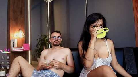 A video of a shirtless, bearded man with glasses and a woman with long black hair drinking from a yellow cup, both wearing casual clothes.