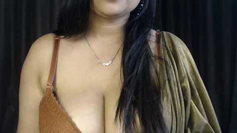 Video of a woman with long, straight black hair, wearing a brown lace bra and a draped olive-green fabric, against a dark background, showcasing her cleavage.