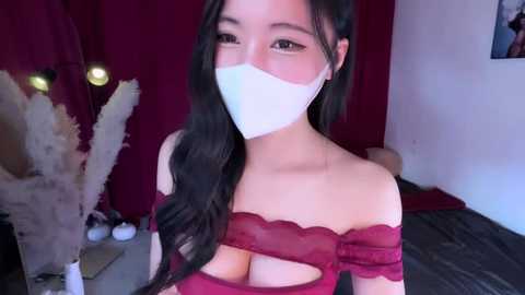 Video of an Asian woman with long black hair and light skin, wearing a red lace off-shoulder top revealing cleavage, white mask, and red curtains in the background.