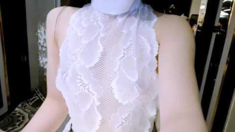 Video of a slender, fair-skinned woman wearing a white, sheer lace top with floral patterns, paired with a light blue turtleneck. The background shows a dimly lit, modern room with glass doors and a patterned rug.