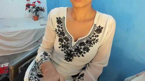 Media: Video of a light-skinned woman with a medium build, wearing a white long-sleeve dress adorned with intricate black embroidery, seated in a chair against a blue wall. The background includes a table with red flowers and a white cloth.