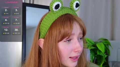 Video of a young woman with long red hair wearing a green crocheted frog hat with googly eyes, smiling. Background includes a green plant, a computer monitor, and a mirror.