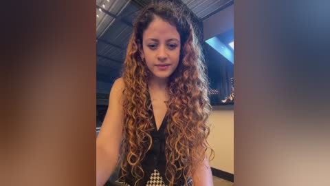 Video of a young woman with long, curly, golden-brown hair, wearing a black sleeveless top, standing indoors with industrial-style ceiling and dim lighting.