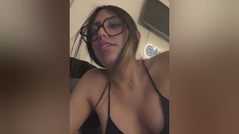 Video of a woman with medium skin tone, wearing black lingerie and glasses, smiling slightly, with a blurred background featuring a flat-screen TV and wall decor.