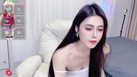 Video of an Asian woman with long black hair, fair skin, and red lipstick, wearing a white off-shoulder top, seated on a white leather chair. Background includes a gray bed and a white wall with a colorful light display.