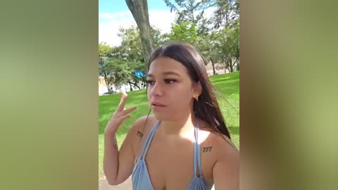 Video of a young woman with long dark hair, wearing a light blue halter top, standing in a lush green park. She gestures with her hand and has tattoos on her shoulders.