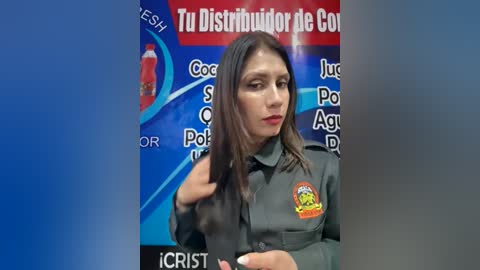 Video of a woman with long dark hair, wearing a gray uniform with a badge, standing in front of a blue and red promotional poster for a \"Cocaine\" show.