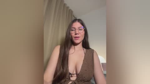 A video of a middle-aged woman with long brown hair, wearing glasses and a brown sleeveless top, standing indoors against beige walls and curtains.