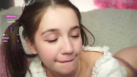 Video of a young, fair-skinned girl with brown pigtails, wearing a white, frilly dress, with closed eyes and a slight smile. Background features blurred pink and beige tones.