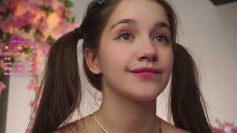 Video of a young, light-skinned girl with pigtails, smiling, wearing a pink top, against a blurred background of pink flowers.