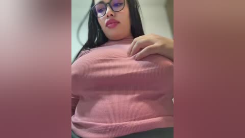 Video of a plus-sized Latina woman with long black hair, wearing glasses, a pink ribbed turtleneck, and a green skirt, standing in a dimly lit room.