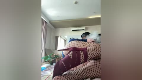 A video of a woman with short black hair, wearing fishnet stockings and a burgundy bra, lying on a bed in a dimly lit, beige room with a window, a potted plant, and an air conditioner.