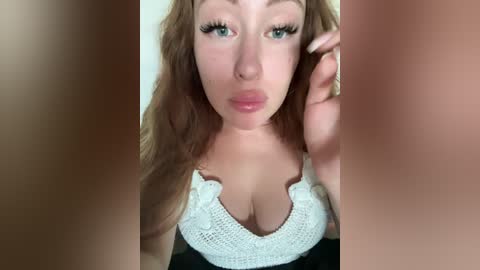Video of a young, fair-skinned woman with long, wavy red hair, wearing a white, lace-trimmed top that reveals cleavage. She has full lips, arched eyebrows, and long lashes. The background is blurred, focusing on her face.