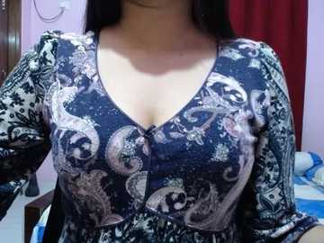 Video of a woman with long black hair wearing a dark, patterned, V-neck blouse with floral designs and a plunging neckline, partially visible cleavage. Background includes light pink walls, a white door, and a red curtain.