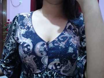 Video of a woman's upper body in a deep blue, patterned blouse with a plunging V-neckline, her right hand raised to her face, partially visible, in a dimly lit room with pink walls and a wooden door.