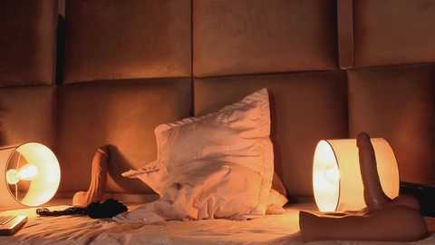 Video of a cozy bedroom scene featuring a plush white pillow, two lit lamps, and a wooden figure with outstretched arms on a beige bed, bathed in warm, inviting light.