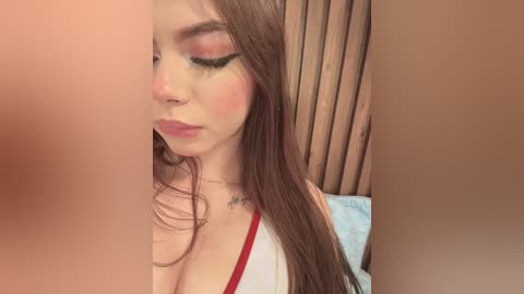 Video of a young Caucasian woman with long brown hair, wearing dramatic makeup, including heavy eyeliner and blush, and a white top with red trim, against a brown curtain backdrop.