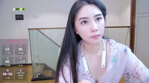 Video of an Asian woman with long black hair, wearing a white floral shirt, sitting at a table in a modern room with a glass staircase in the background.