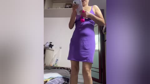 Video of a light-skinned woman in a tight, sleeveless, purple mini dress, holding a smartphone, taking a mirror selfie in a cluttered bedroom with a messy bed and a door frame.
