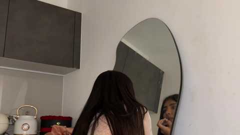 A video of a woman with long, straight, dark hair standing in a modern kitchen, facing a large oval mirror. She's wearing a beige top, and the background features sleek, gray cabinets and a stainless steel kettle on a white countertop.